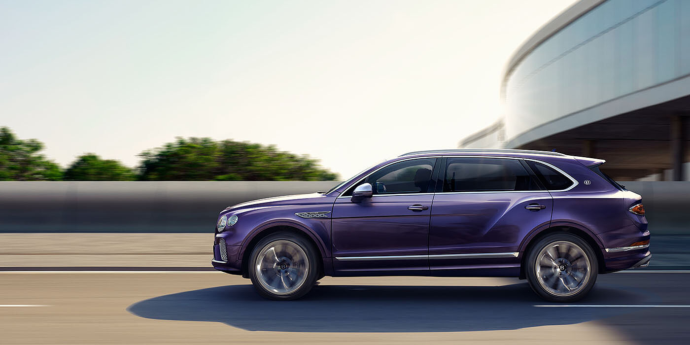 Bentley Cairo Bentley Bentayga Extended Wheelbase Mulliner luxury SUV profile in Tanzanite Purple paint, driving on an country road past trees.