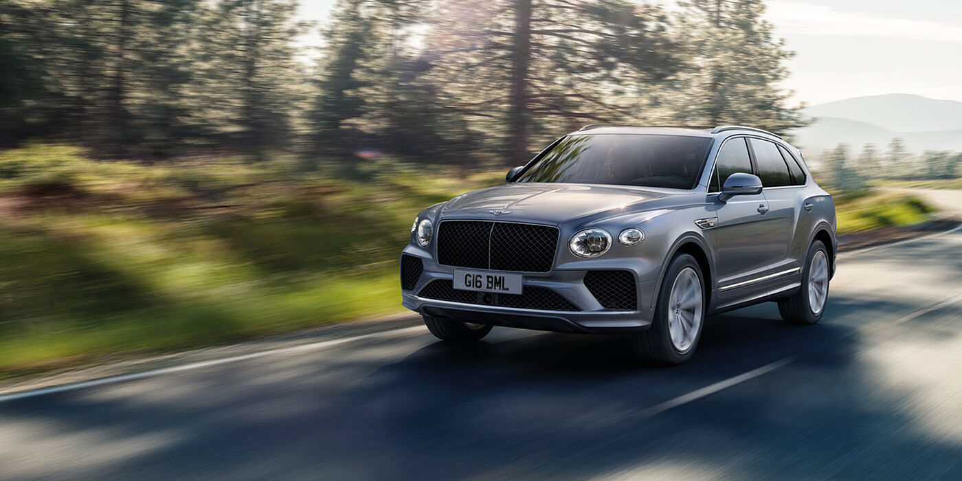 Bentley Cairo Bentley Bentayga luxury SUV front three quarter in Moonbeam paint, driving on an country road past trees.