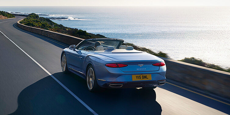 Bentley Cairo New Bentley Continental GTC Azure convertible rear view in Light Windsor blue paint with 22 inch ten spoke wheels driving dynamically on an ocean road in the sunshine